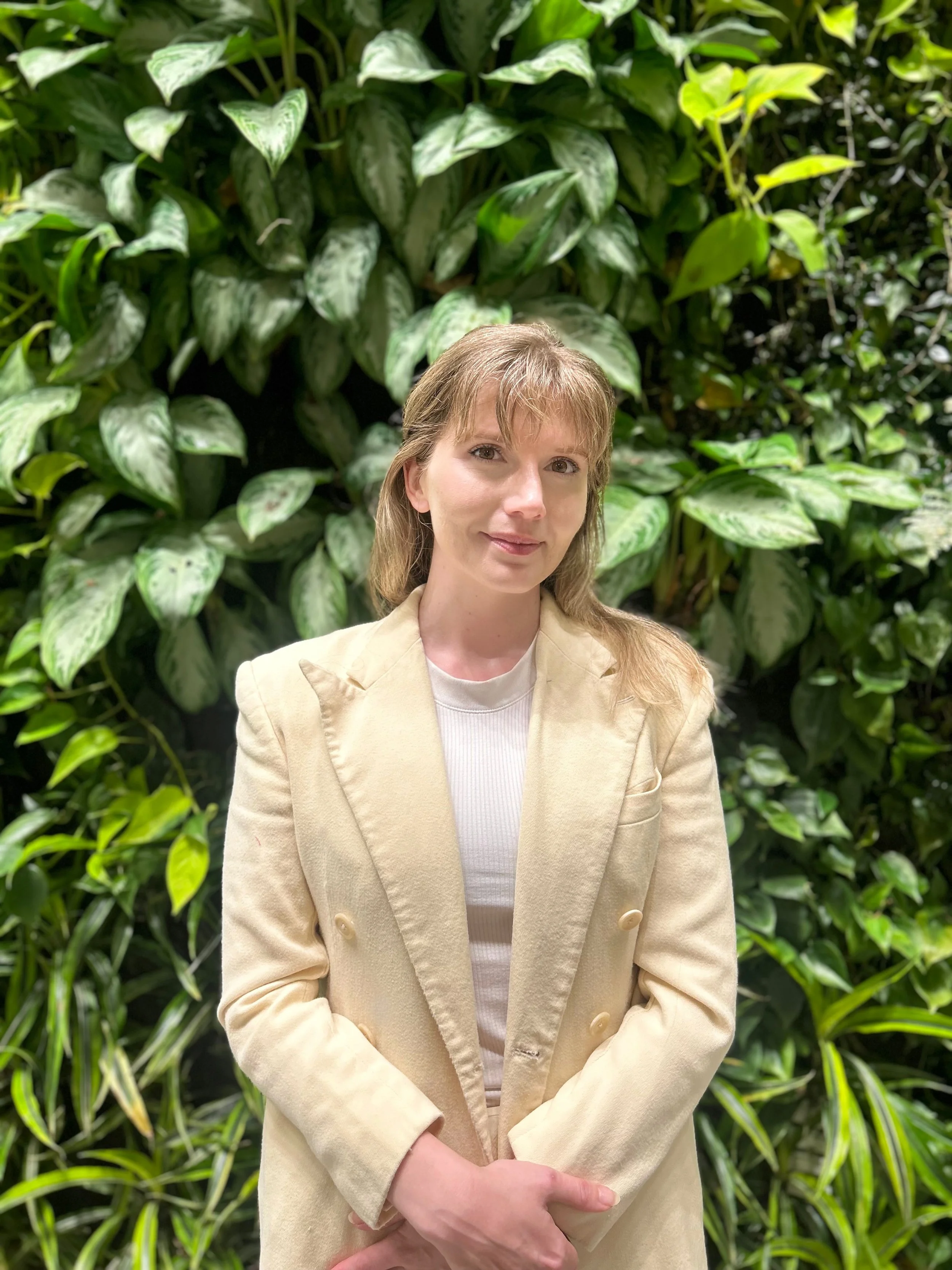 A woman with light brown hair, wearing a beige blazer and white shirt, stands in front of green leafy plants and looks at the camera with a slight smile.
