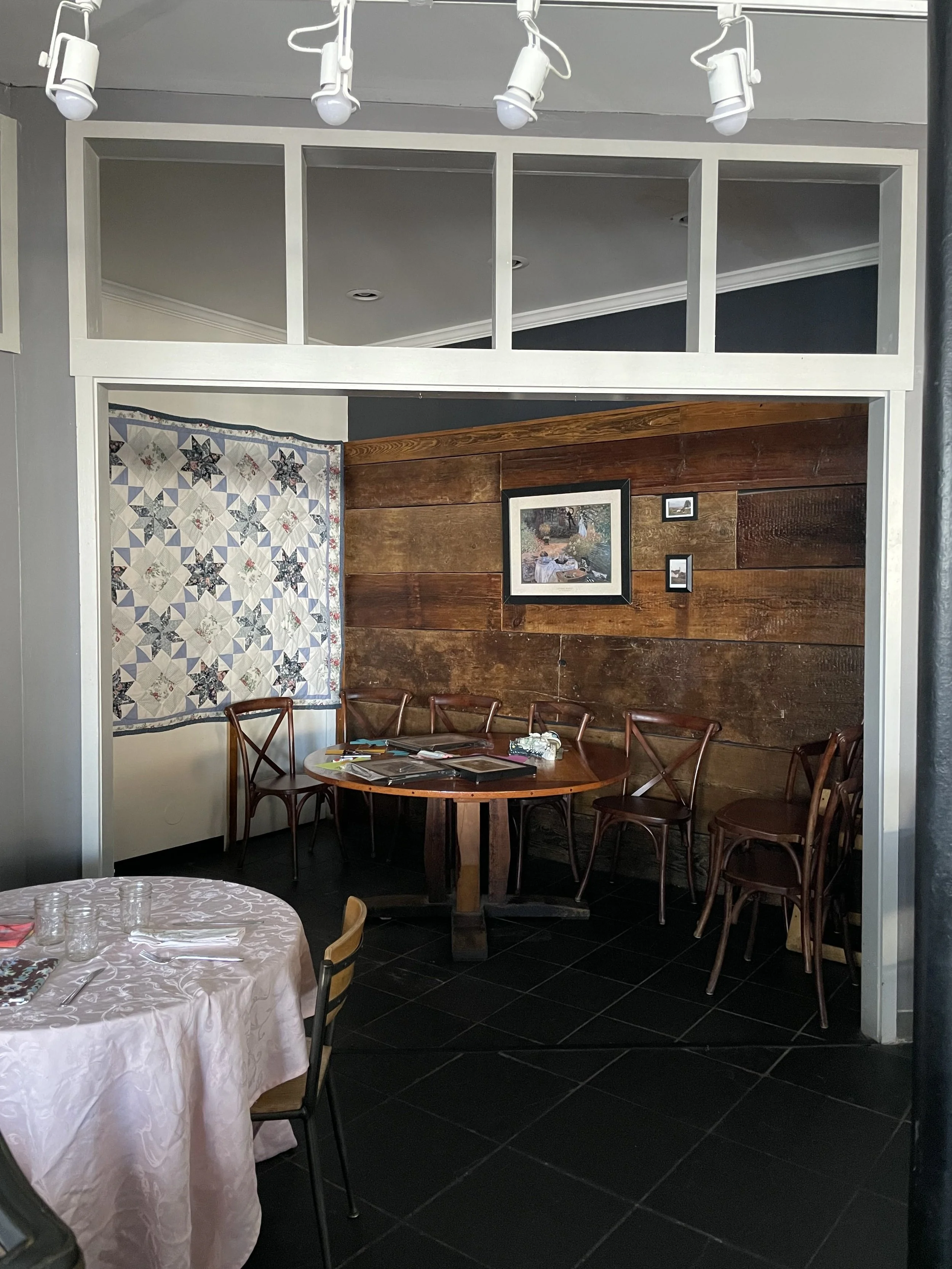A cozy restaurant corner with a wooden wall, a framed painting, and several chairs around a table, with some items on it.