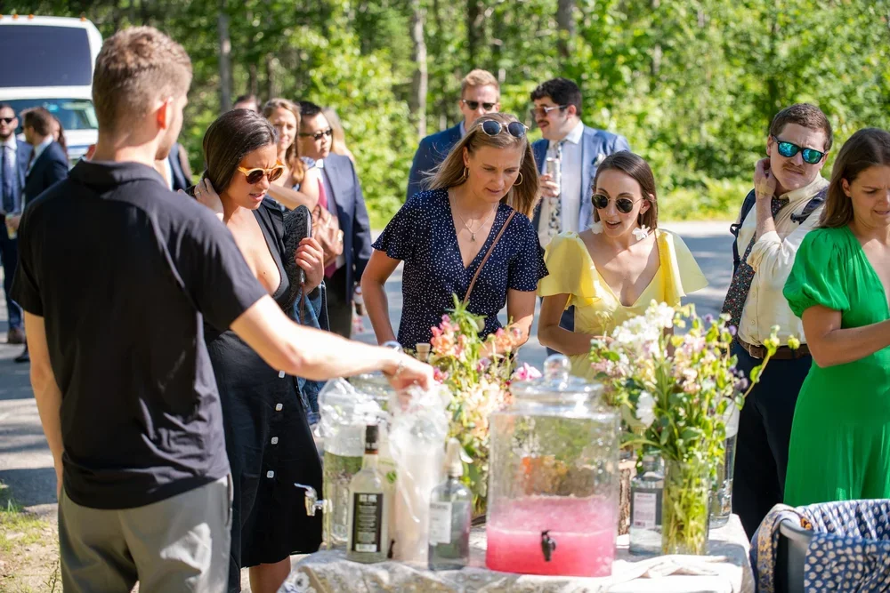 People gathered outdoors around a table with drinks and flowers, chatting in daylight.