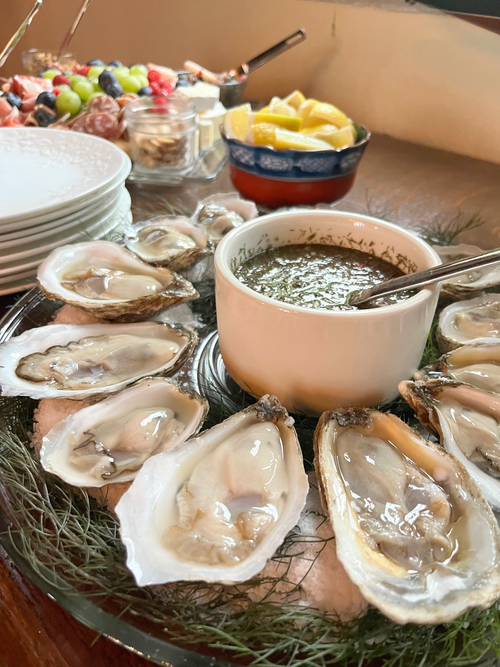 Fresh oysters on the half shell with a cup of green cocktail sauce, lemon wedges, and various fruits and condiments in the background.