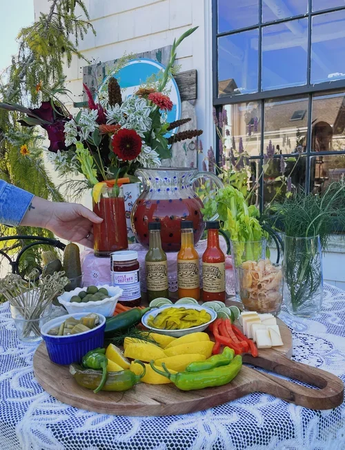 A table with a wooden serving board of sliced bell peppers, cucumbers, and cheese, with condiments, pickles, and hot sauce bottles, surrounded by flowers and a pitcher of red beverage, set against a house with large windows and sky reflections.