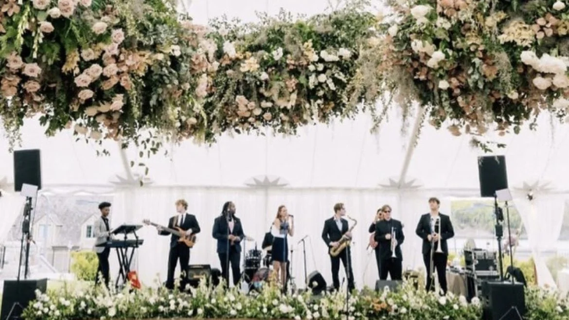 A band performing with seven members, including a keyboardist, guitarist, singer, saxophonist, and other musicians, on a decorated stage with floral arrangements.