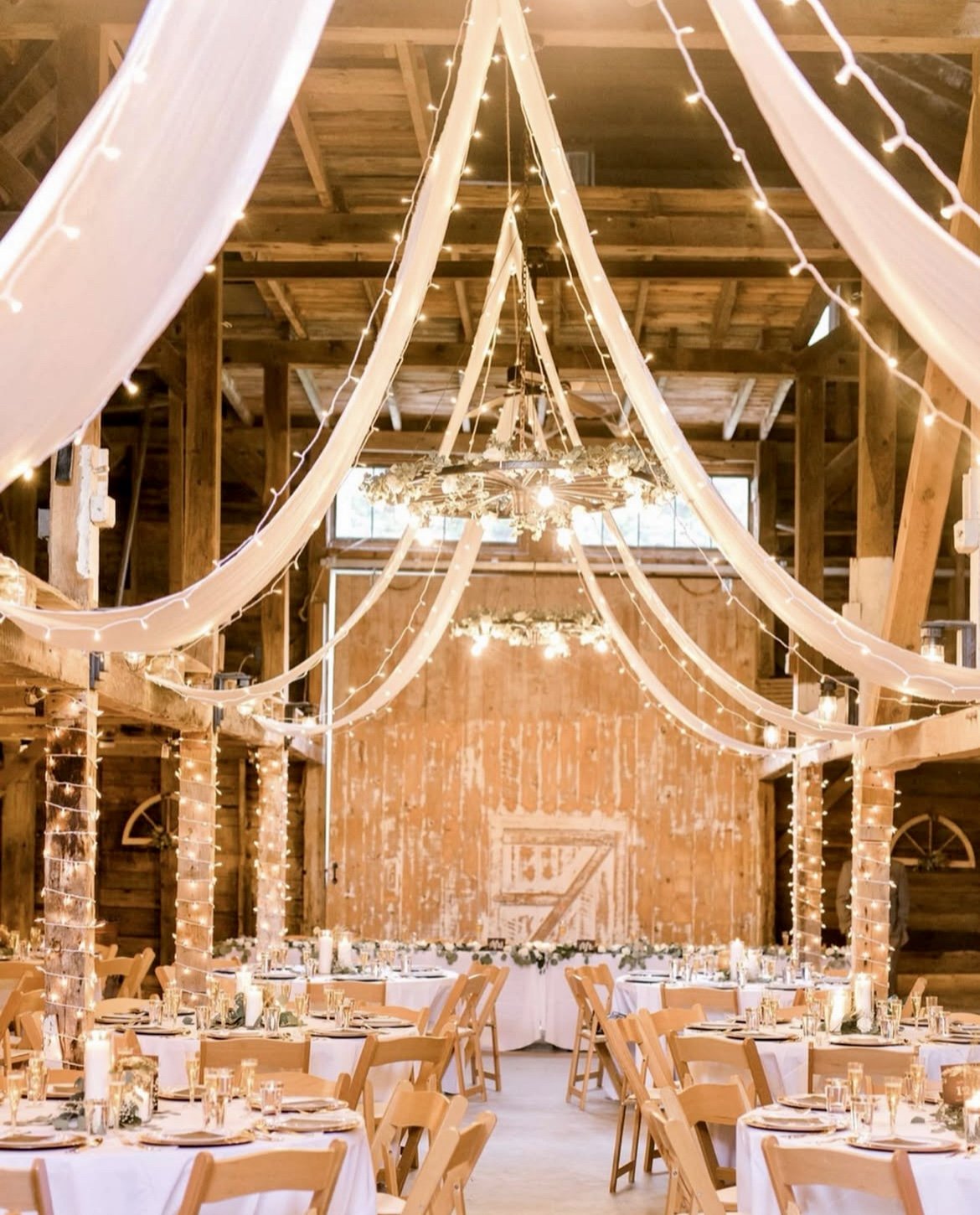 Rustic wedding reception with round tables, white linens, candles, and wooden chairs, decorated with string lights and draped fabric hanging from the ceiling in a barn-style venue.