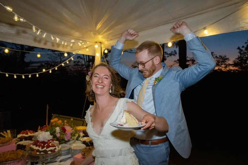 A couple celebrating at a wedding reception, smiling and dancing under string lights with a table of desserts and flowers in the background.