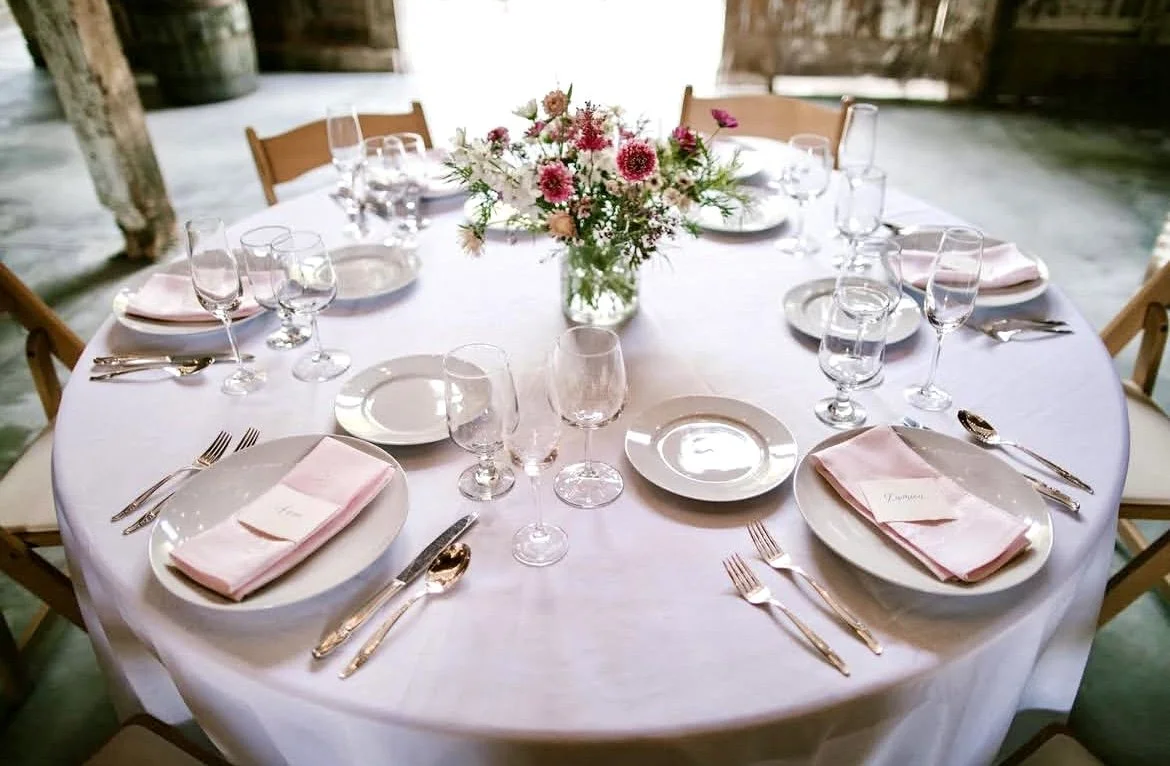 Round banquet table set for a formal event with pink napkins, white plates, various glasses, silverware, and a floral centerpiece in a rustic venue.