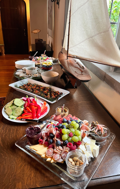 A wooden table set with a sailboat model and an assortment of food including a cheese and fruit platter, sliced cucumbers and red bell peppers, assorted dips and spreads, and candies, with a window and hallway in the background.