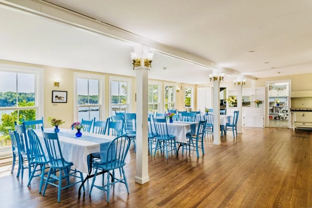 Bright dining area with several tables covered with white tablecloths and adorned with small flower arrangements in blue vases, surrounded by blue chairs, with large windows showing a scenic outdoor view of water and greenery.