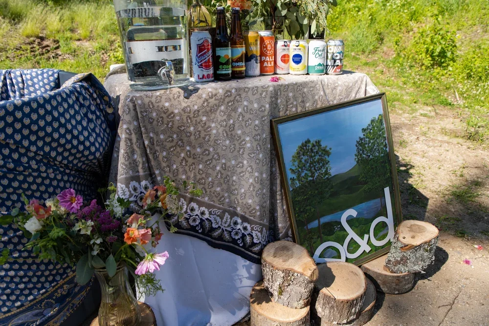 Outdoor table with a floral tablecloth, various bottles and cans of beverages, a large water dispenser, a bouquet of flowers in a vase, and a framed mirror resting against tree stumps on the ground. Green grass and trees are visible in the background