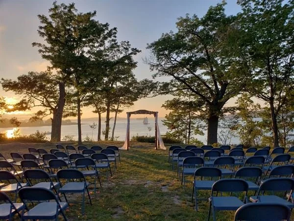 Outdoor wedding ceremony setup with rows of chairs facing a wooden arch on a grassy area by a lake, with trees and a sunset in the background.