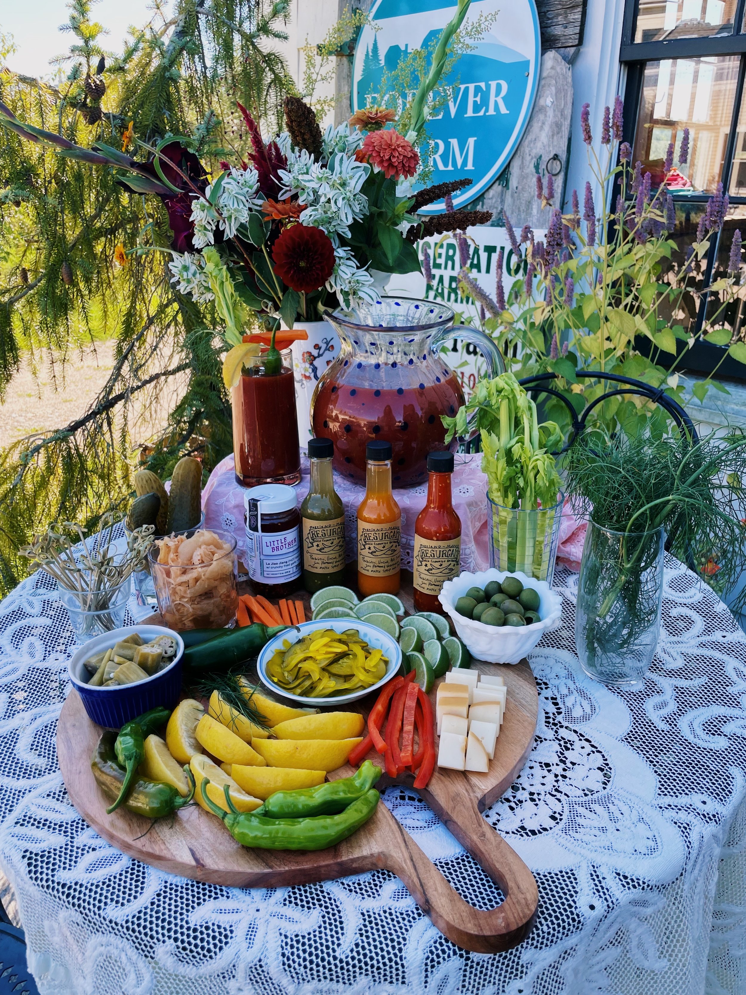 A table set with fresh vegetables, cheeses, lime slices, hot sauce, and condiments, surrounded by plants and flowers outdoors.