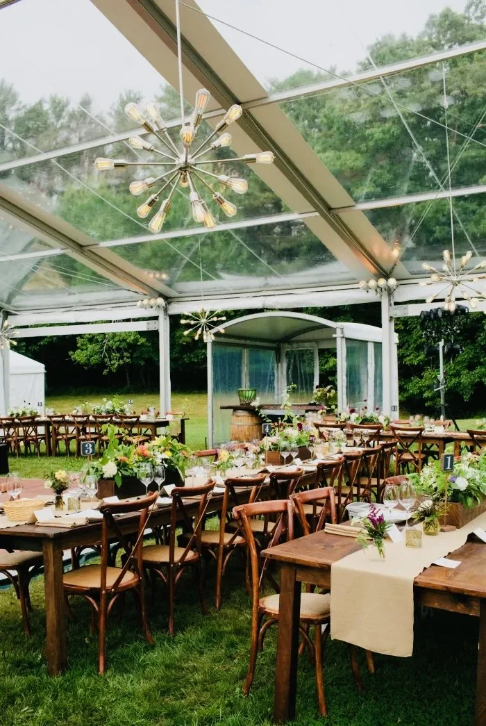 Decorated outdoor event space with wooden tables and chairs, floral centerpieces, and modern chandeliers under a transparent tent ceiling, with a view of green trees and grass outside.