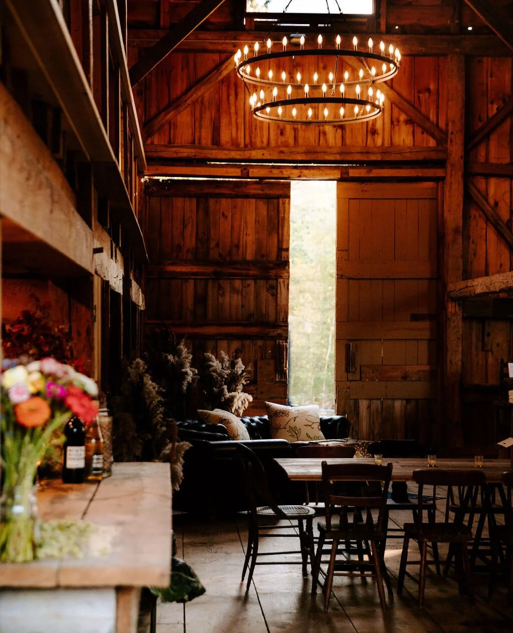 Interior of a rustic barn with wooden walls and ceiling, illuminated by a chandelier with small candles. There are tables and chairs, a leather sofa wreath with pillows, and a bouquet of colorful flowers on a counter.
