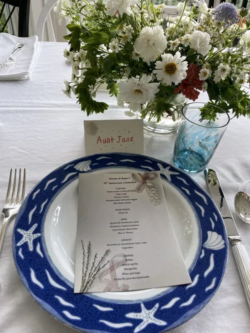 A table setting with a blue fish-and-starfish patterned plate, a printed menu, a bouquet of assorted white and pink flowers in a glass vase, a blue-tinted drinking glass, and a small name card that reads Aunt Jane.