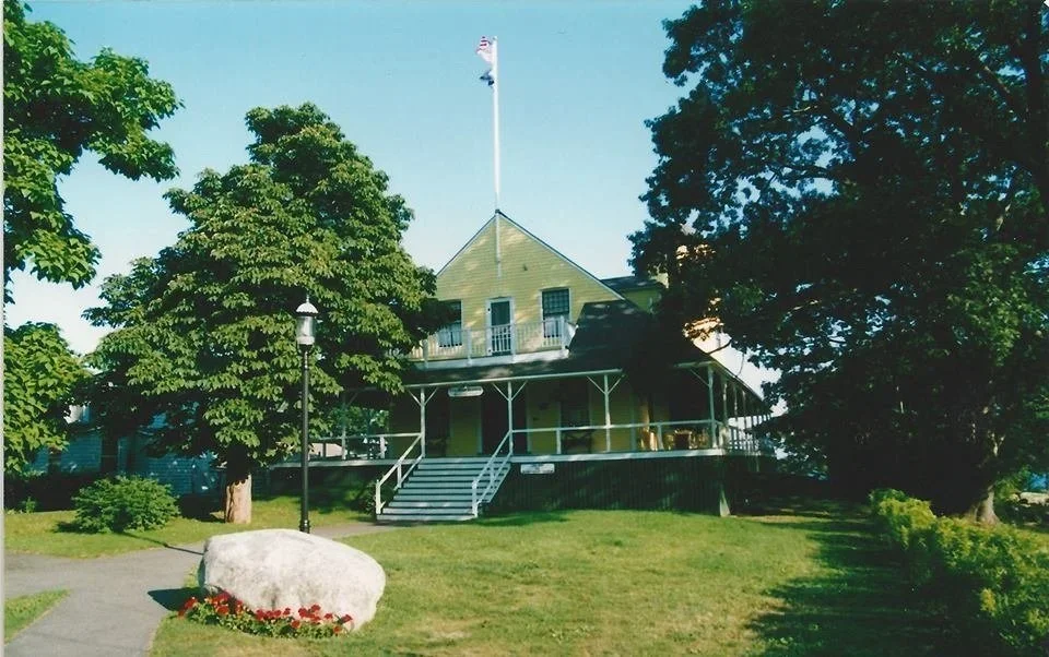 A two-story house with a wraparound porch, a flag on a pole on the roof, surrounded by green trees and grass, with a large rock and flowers in the yard.