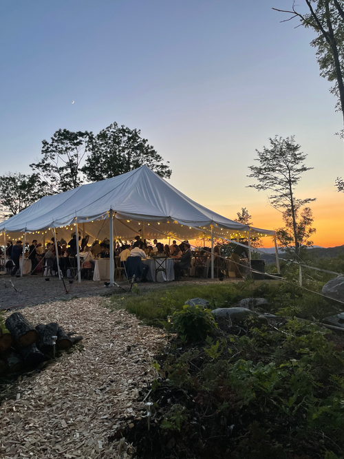 Outdoor sunset outdoor gathering under a large white tent with string lights, people dining, surrounded by trees and nature.