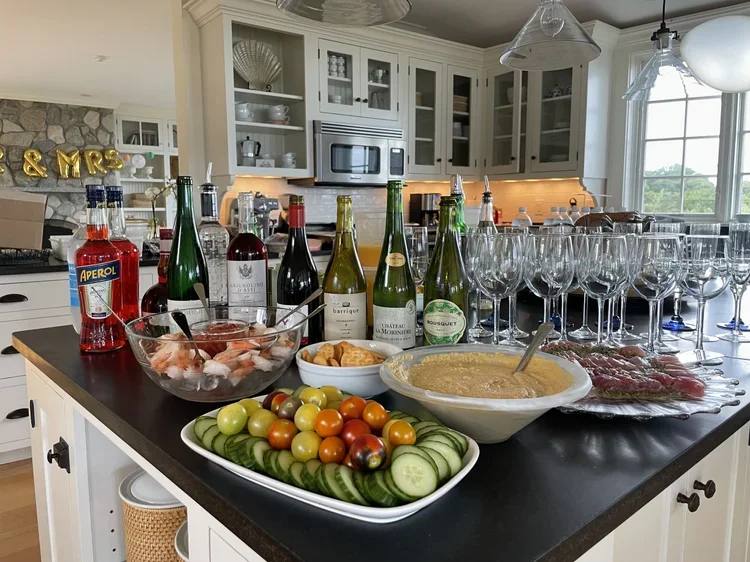 A kitchen island with various appetizers and drinks, including sliced cucumbers and cherry tomatoes, a bowl of dip, raw meat, shrimp, multiple wine bottles, and numerous empty wine glasses.