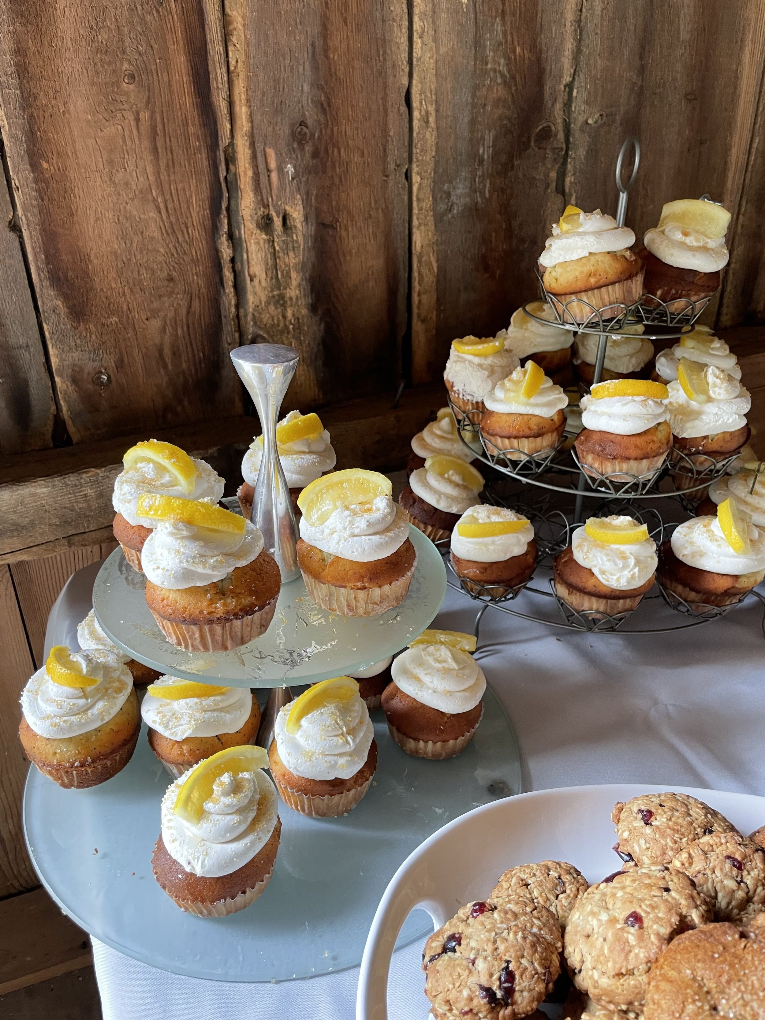 Lemon cupcakes with white frosting and lemon slices, displayed on a tiered serving stand and a plate, with oatmeal cookies with dried fruit on a white tablecloth against a wooden wall background.