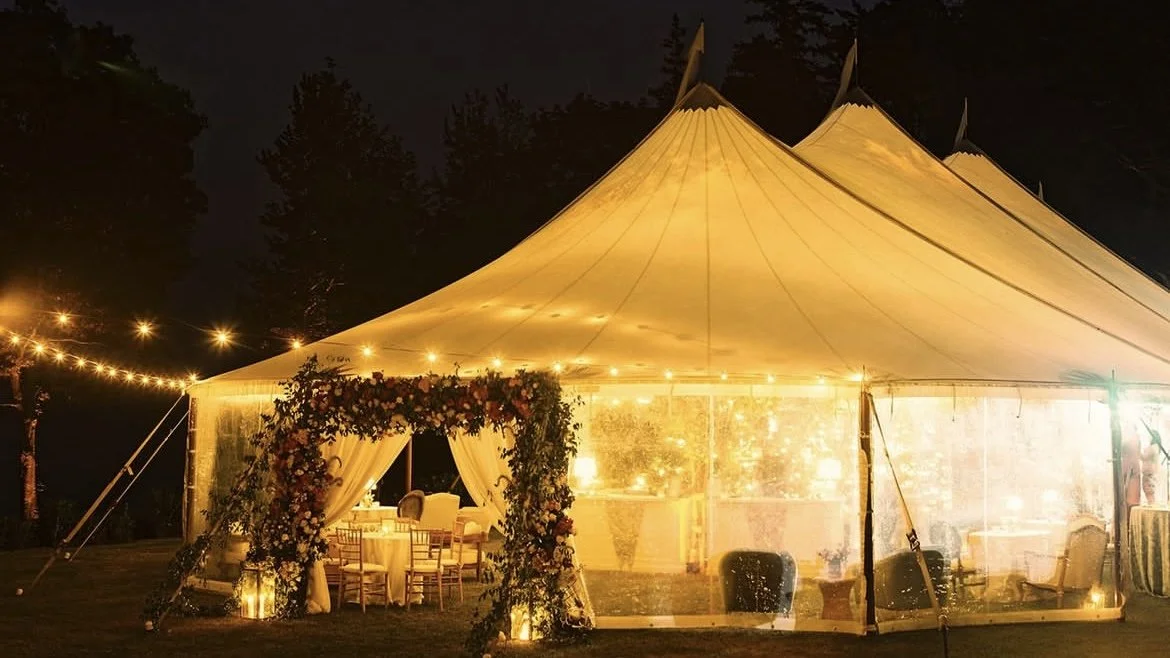 A large illuminated event tent decorated for a celebration at night, with string lights, floral archway entrance, and elegant furniture inside.