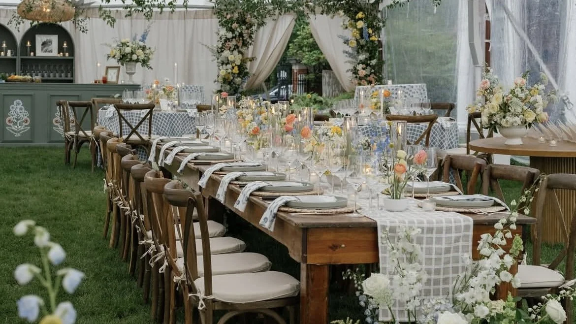 Decorated outdoor dining table with floral centerpieces, candles, and tableware under a canopy.