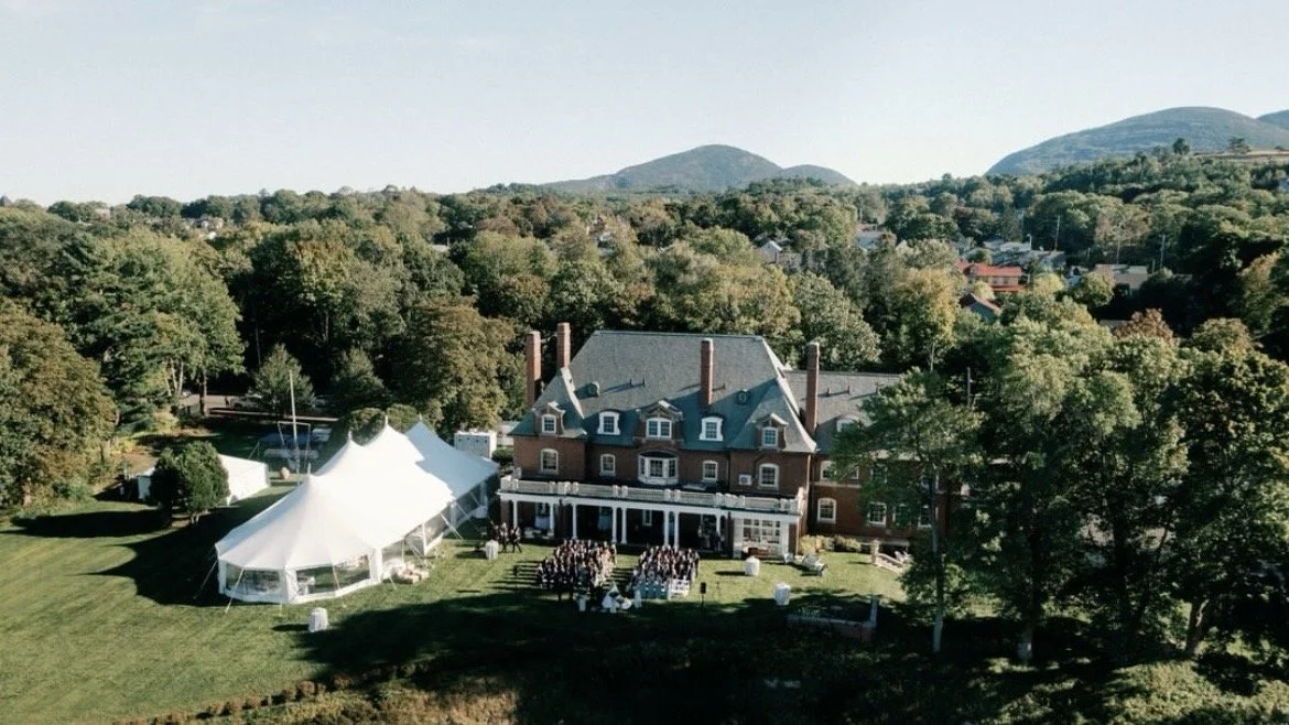 A large outdoor wedding ceremony with guests seated in front of a historic brick mansion, a white event tent nearby, surrounded by trees and hills.