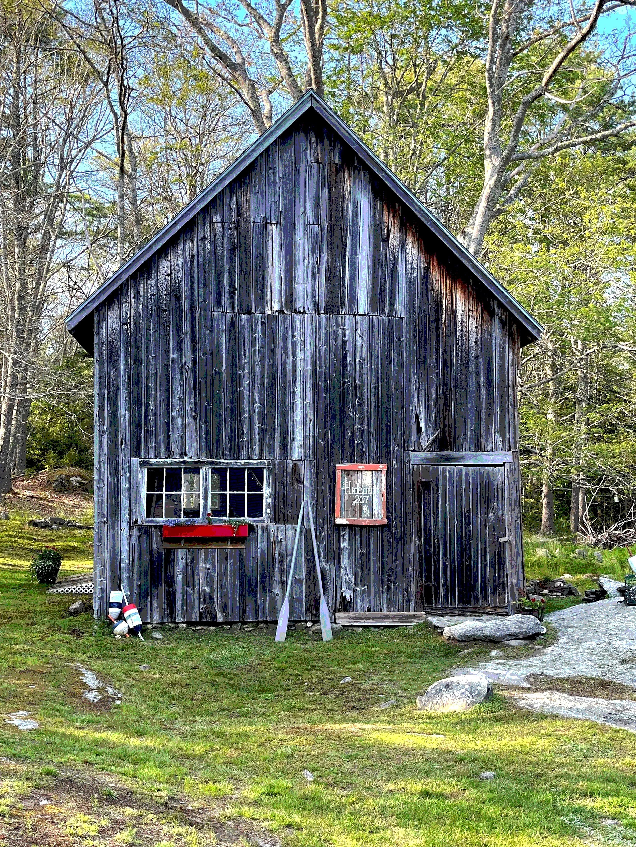 Old weathered black wooden barn in a yard with grass, large rocks, and trees in the background.