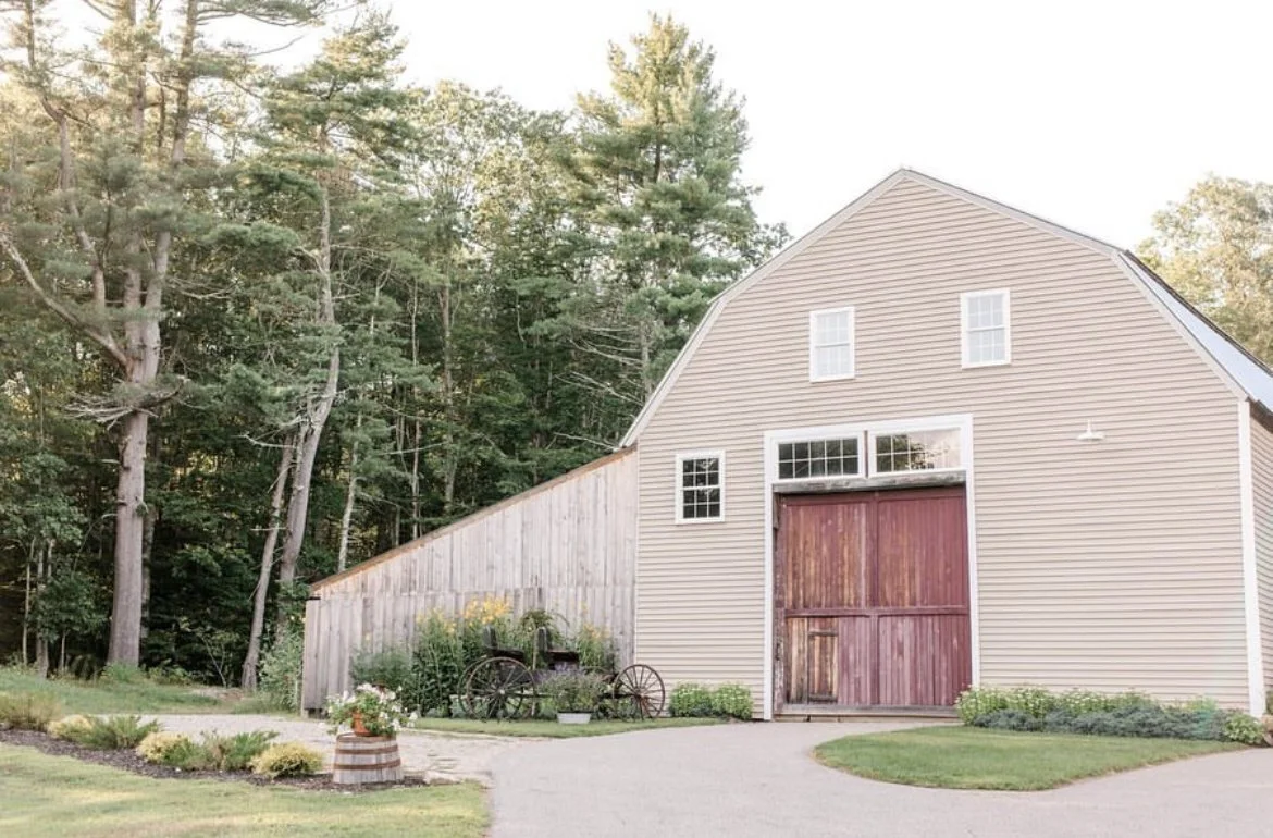 A beige barn with red sliding doors, multiple windows, surrounded by a landscaped yard with flower pots, a wagon, and tall trees in the background