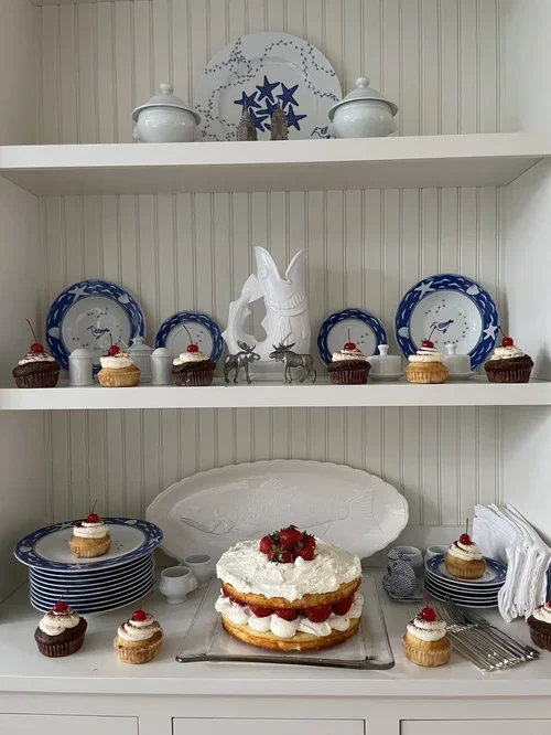 White dining hutch decorated with blue and white dishware and cupcakes, featuring a strawberry shortcake on a glass platter in the center.