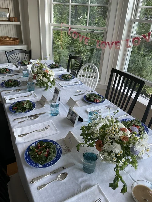 A decorated dining table set for a meal with floral centerpieces, salad plates, water glasses, and place cards, near a window with a pink 'Love' banner hanging in the background.