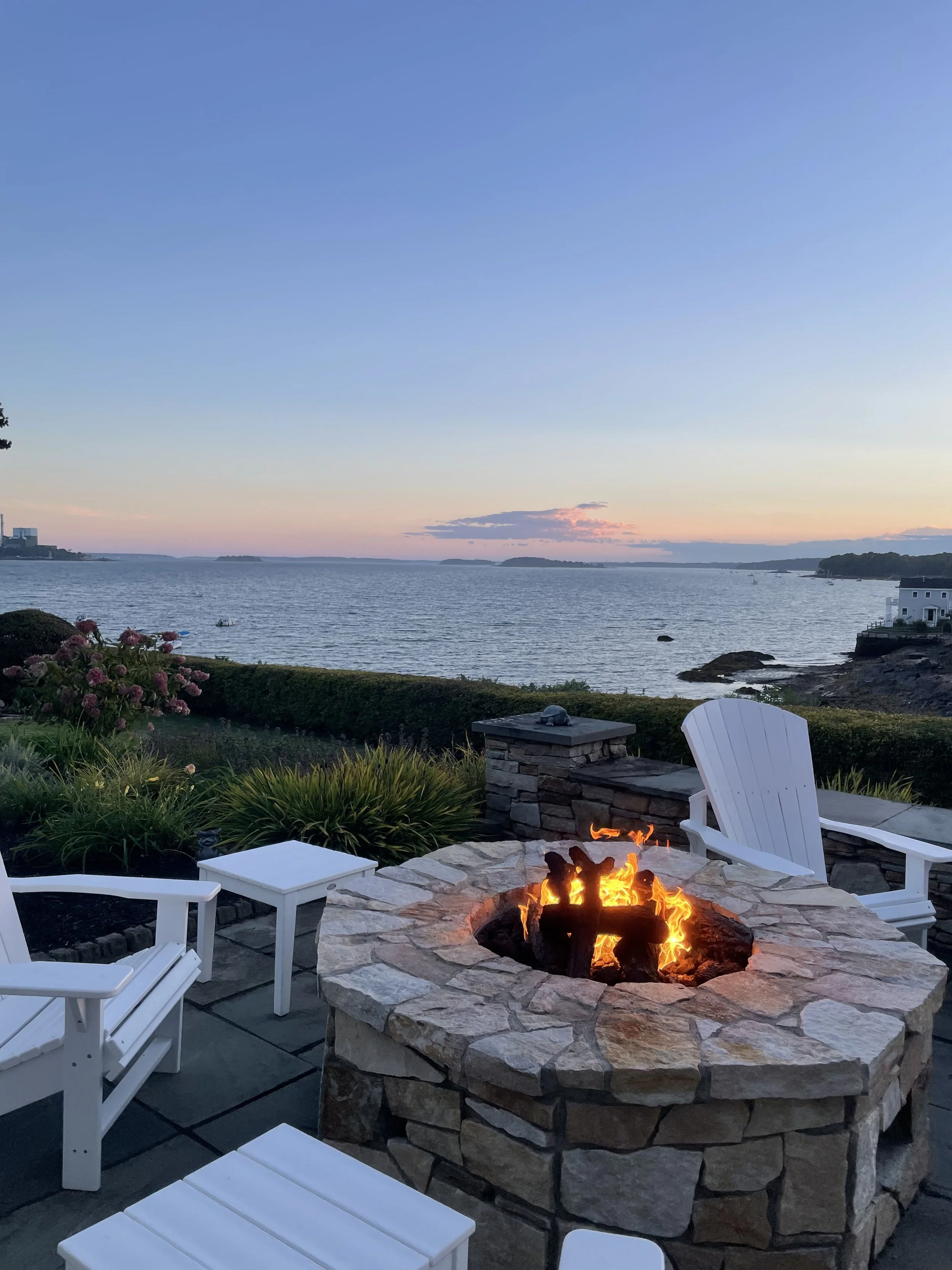 A scenic outdoor patio with white chairs and a stone firepit overlooking a body of water during sunset.