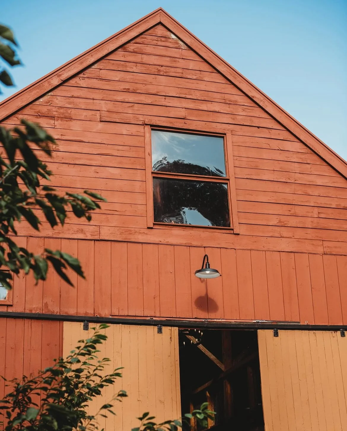 A two-story wooden barn with red-painted siding, a large window on the upper level, and sliding barn doors on the lower level, with some greenery in the foreground and a clear blue sky.