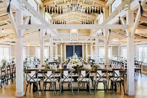A spacious, well-lit event hall with wooden beams, decorated for a celebration, with round tables set with flowers and chairs, and an upper balcony with hanging decorations.