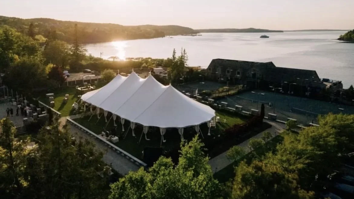 An outdoor event tent on a lawn overlooking a body of water at sunset, with trees and mountains in the background.