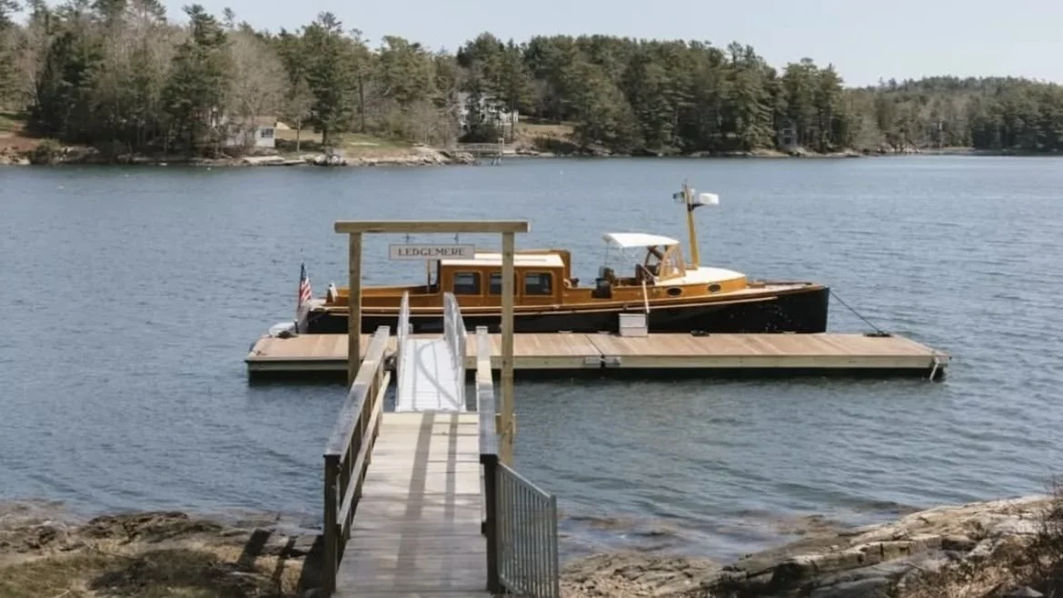A wooden dock with a slide extends into a body of water, with a vintage-style boat docked at the end, and trees lining the distant shore.