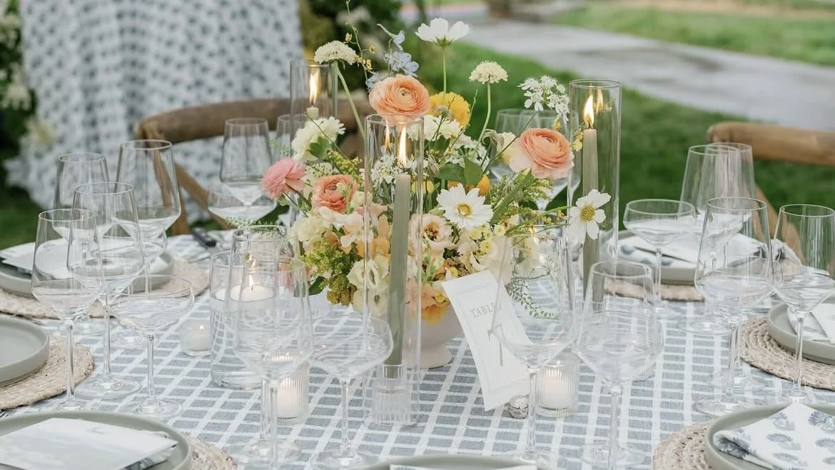 A decorated outdoor table with a floral centerpiece, multiple wine glasses, candles, and place settings for a gathering or wedding reception.