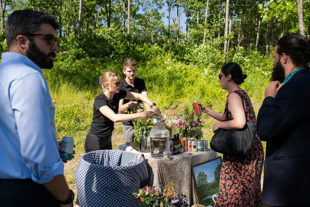 Group of people gathered outdoors around a table with drinks and snacks, during daytime. The background features lush green trees and sunlight.