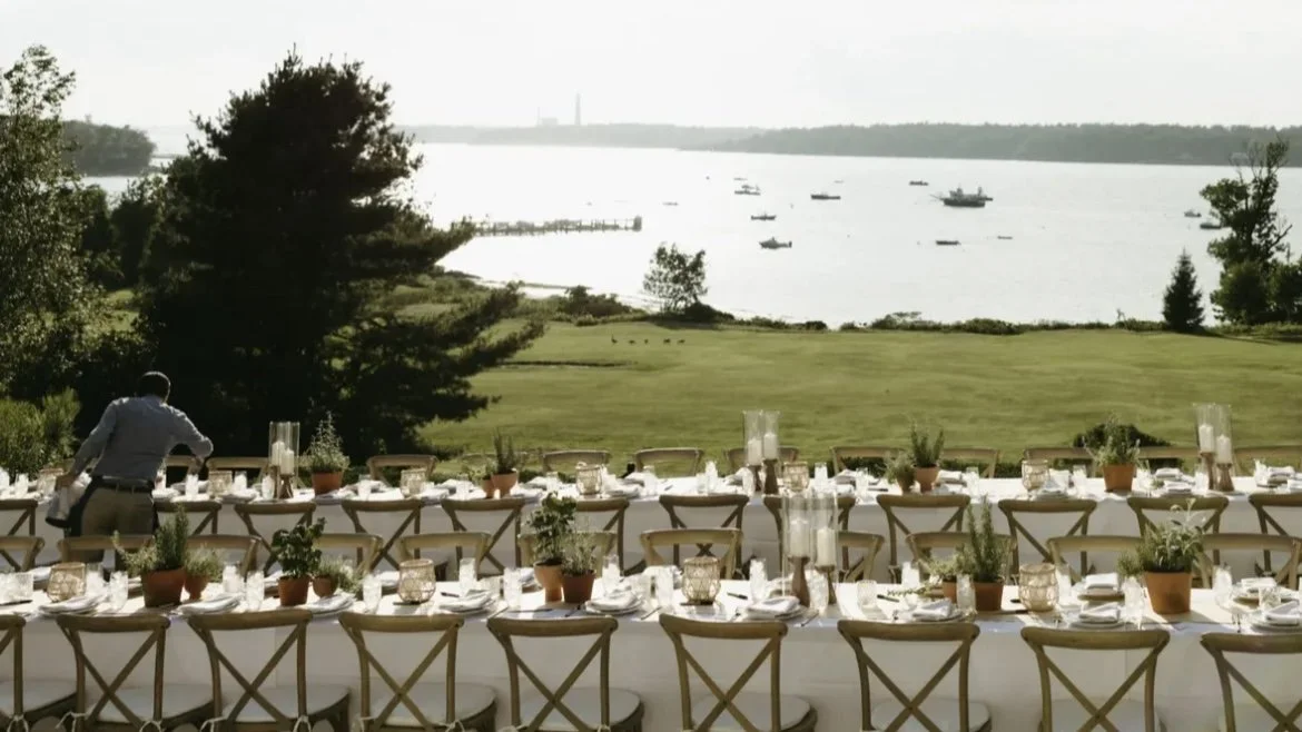 Long outdoor dining table set up with white tablecloths, candles, potted plants, and chairs, overlooking a grassy area, trees, and a body of water with boats, in a scenic outdoor setting.
