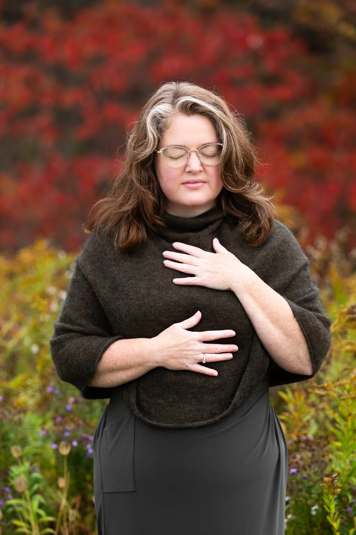 Woman standing outdoors with eyes closed, hands resting on chest and abdomen.