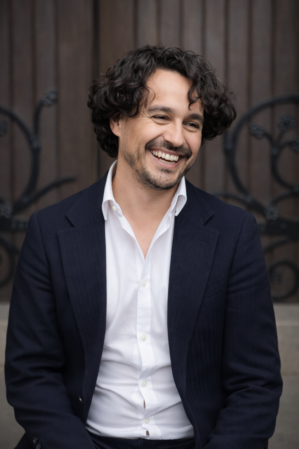 A man with curly dark hair and a beard, wearing a white dress shirt and a dark blazer, smiling outdoors in front of a wooden fence with decorative black wrought iron.