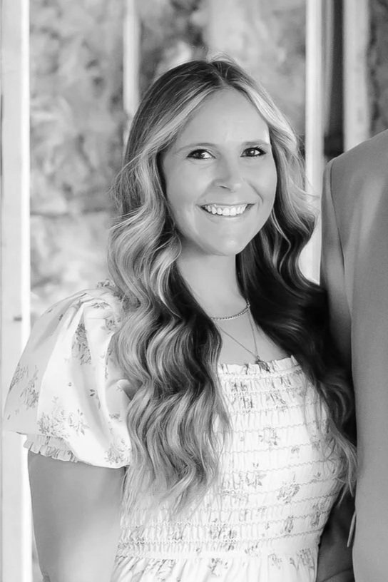 Black and white portrait of a woman with long, wavy hair, smiling and wearing a dark top, with a plant in the background.