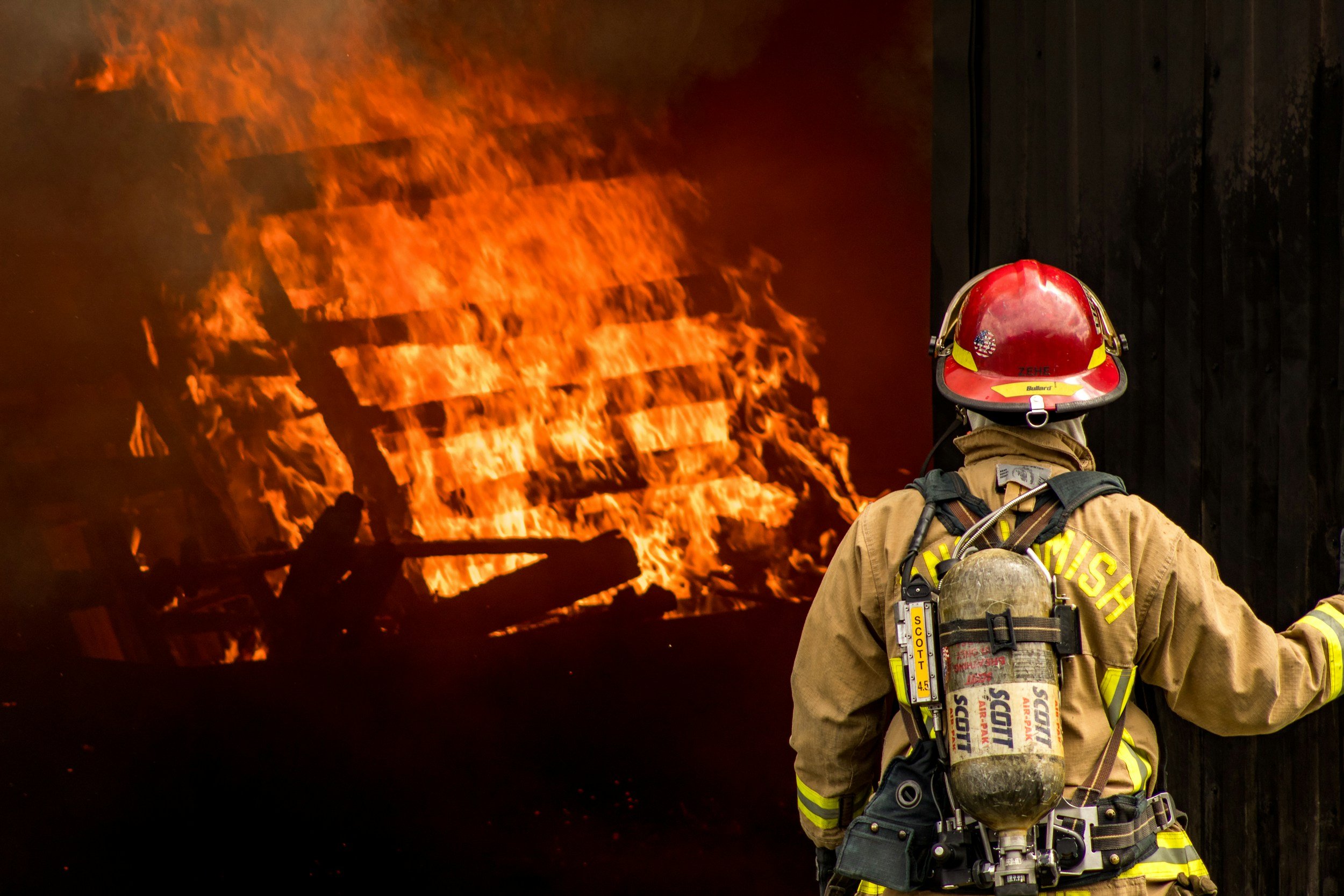 Firefighter wearing protective gear and helmet standing in front of a burning building with flames and smoke.