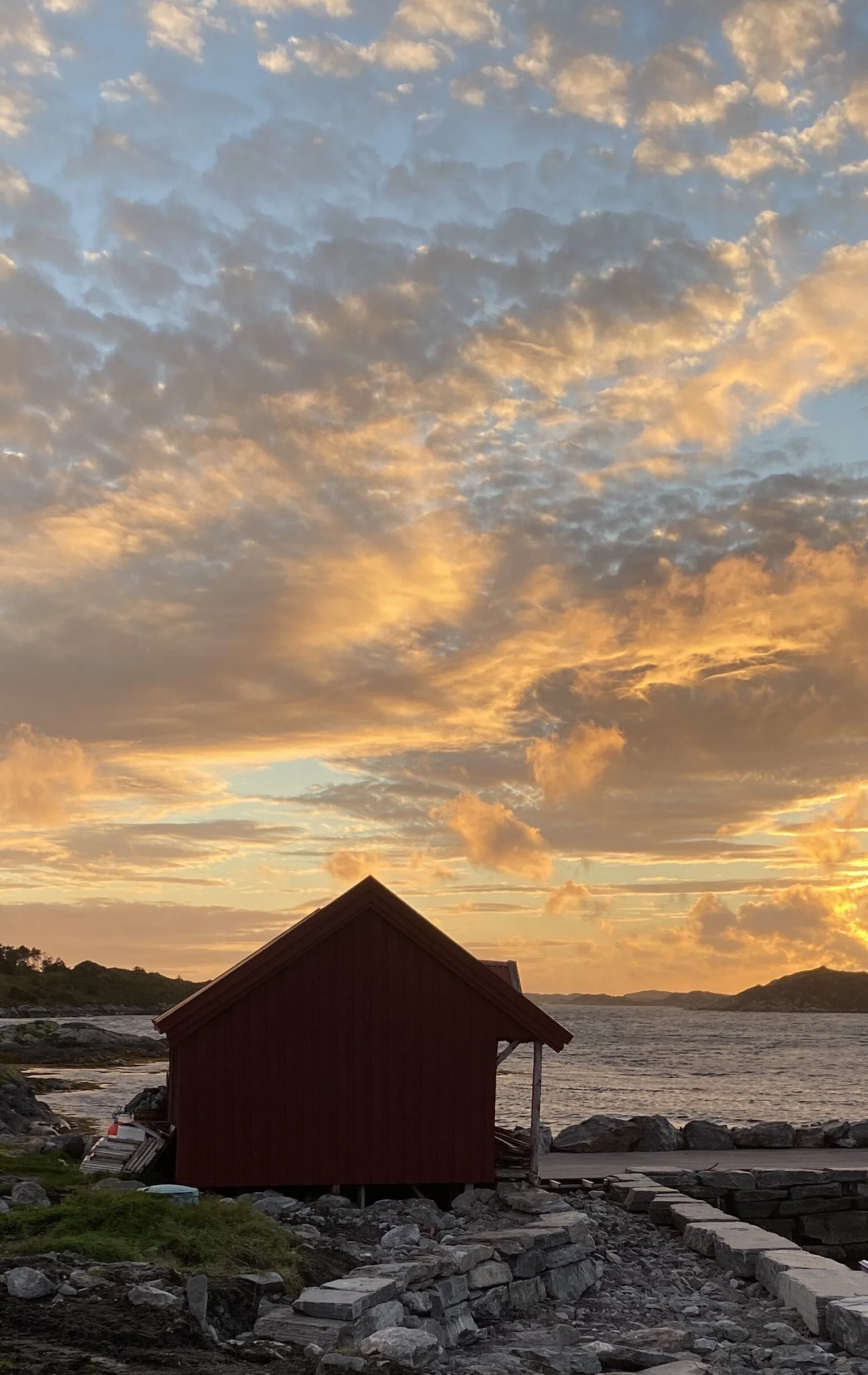 En liten rød hytte ved sjøen under en fargerik solnedgang med skydekke.