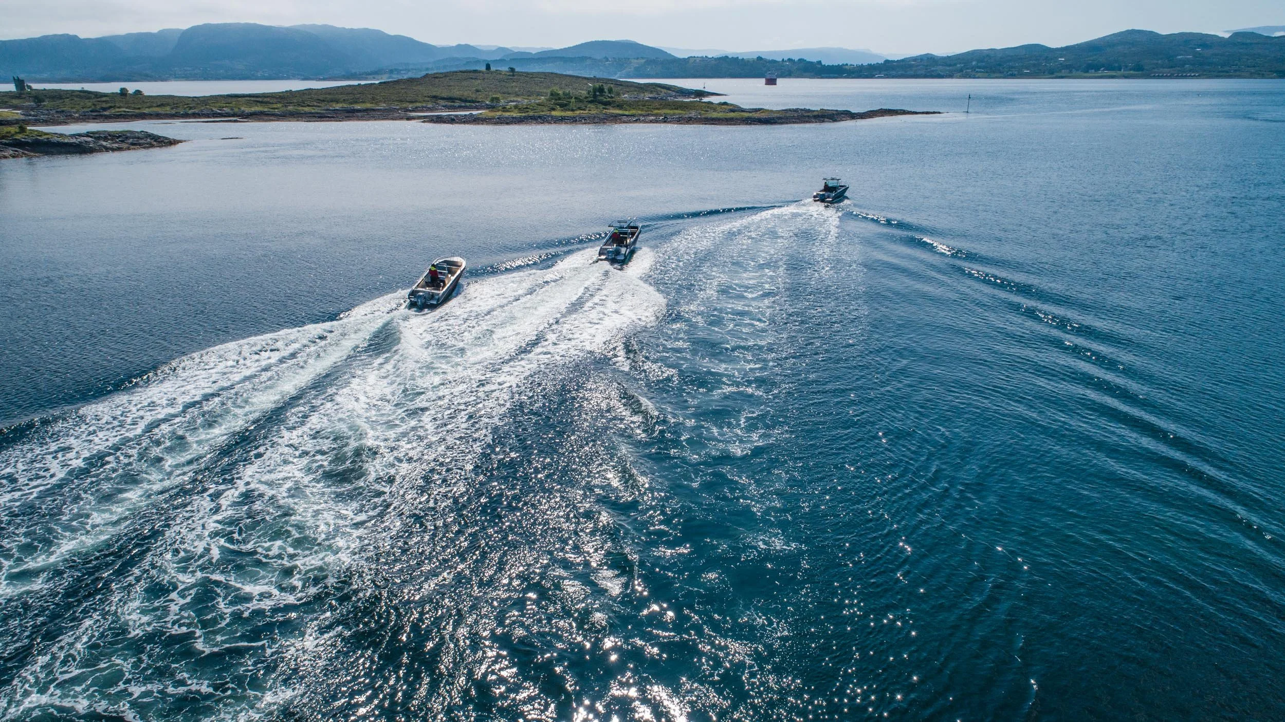 Tre små båter som kjører i vannet, i en fjord med land og fjell i bakgrunnen