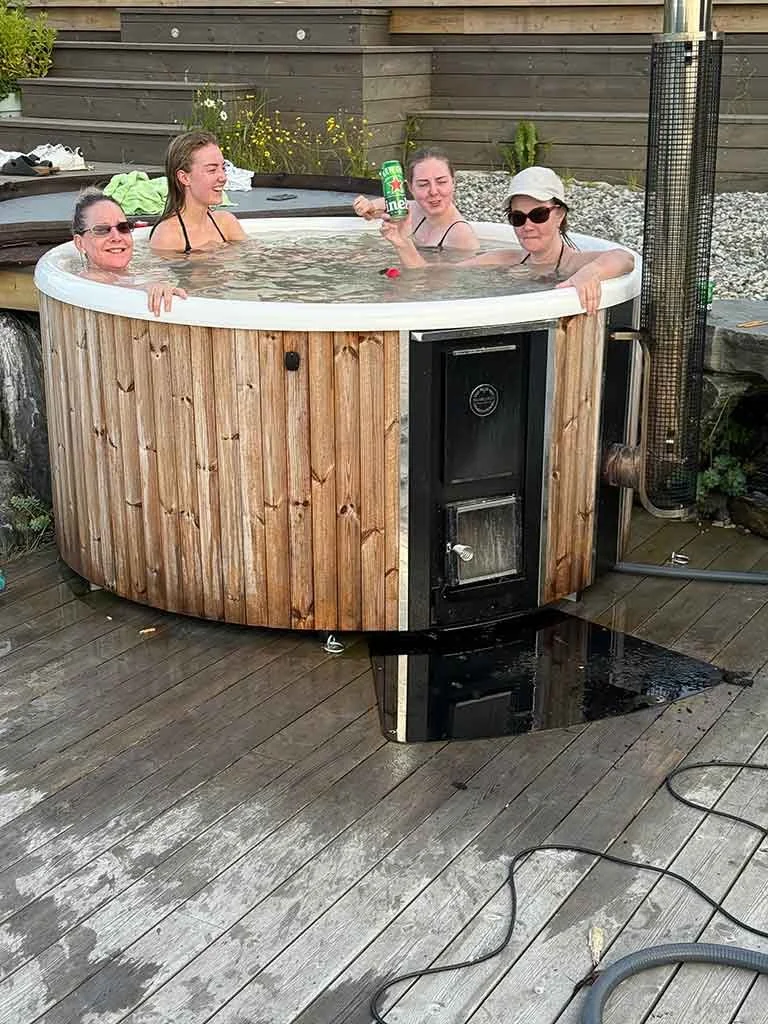 Fire kvinner i et utendørs boblebad på treplatting, en av kvinnene holder en grønn eske. Alle ser ut til å ha det gøy.