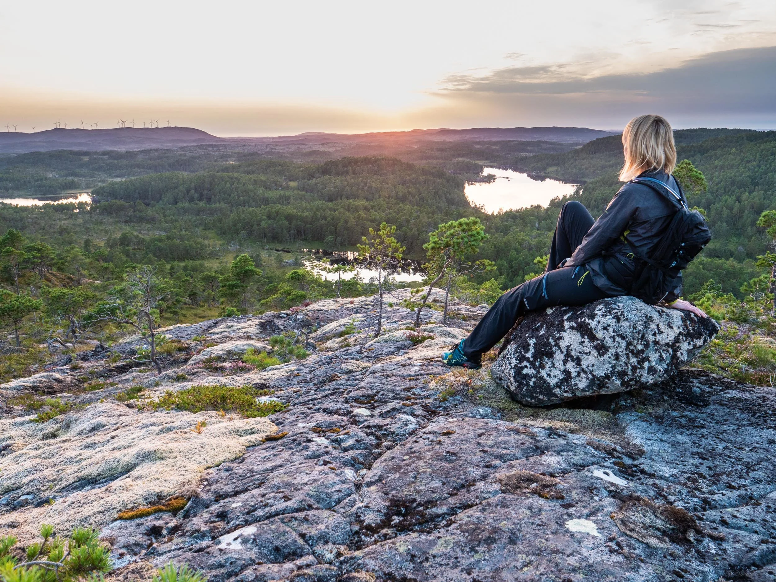 En kvinne sitter på en stein på en fjellknaus og ser utover en grønn skog med innsjøer og fjell i horisonten ved solnedgang.