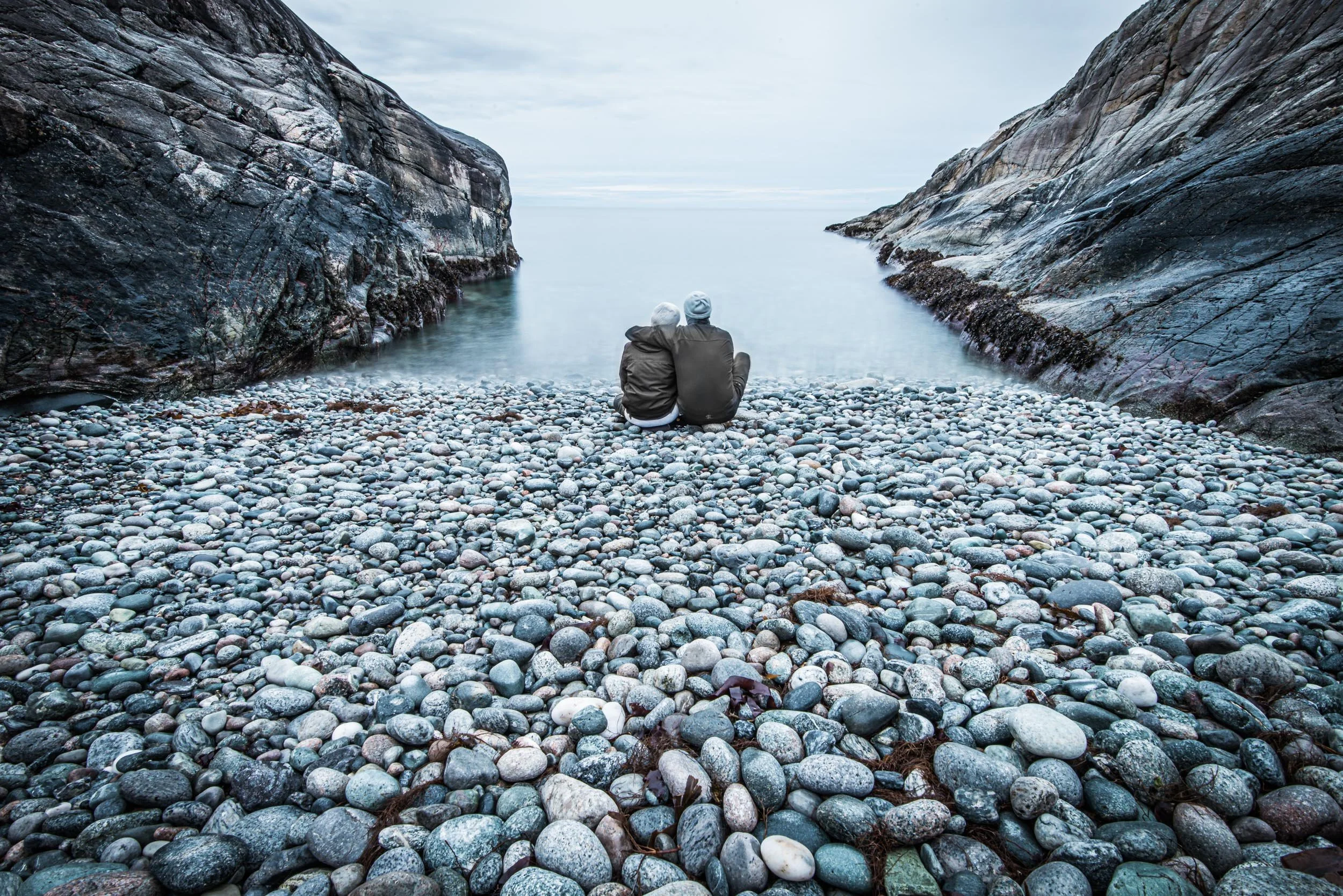 To personer sitter på en steinstrand og observerer vann mellom to klippeformasjoner.