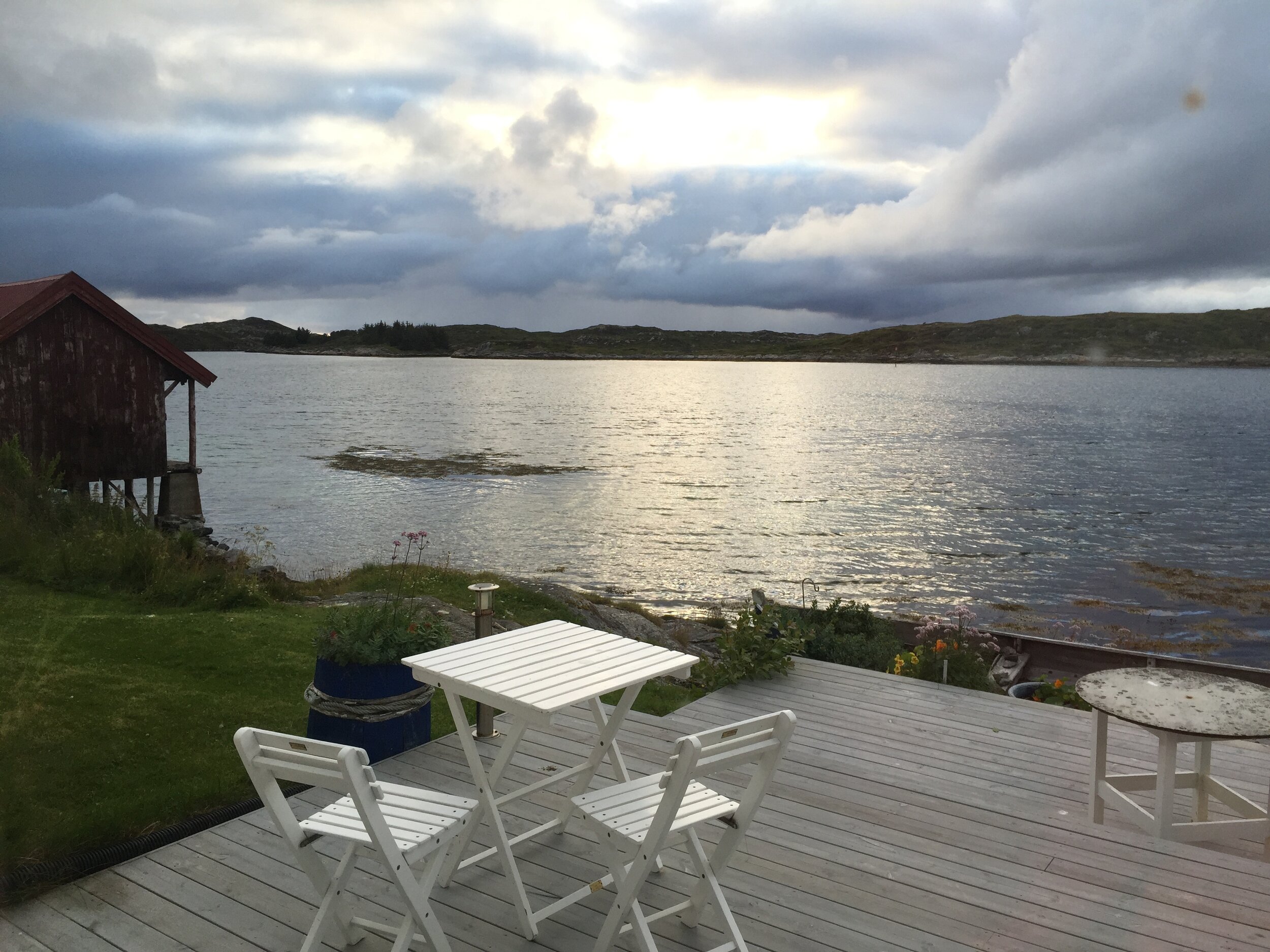 Utsikt over en rolig innsjø med en liten terrasse med hvite møbler foran, en rød låve, og en dramatisk skydomin med svake solstråler i bakgrunnen.
