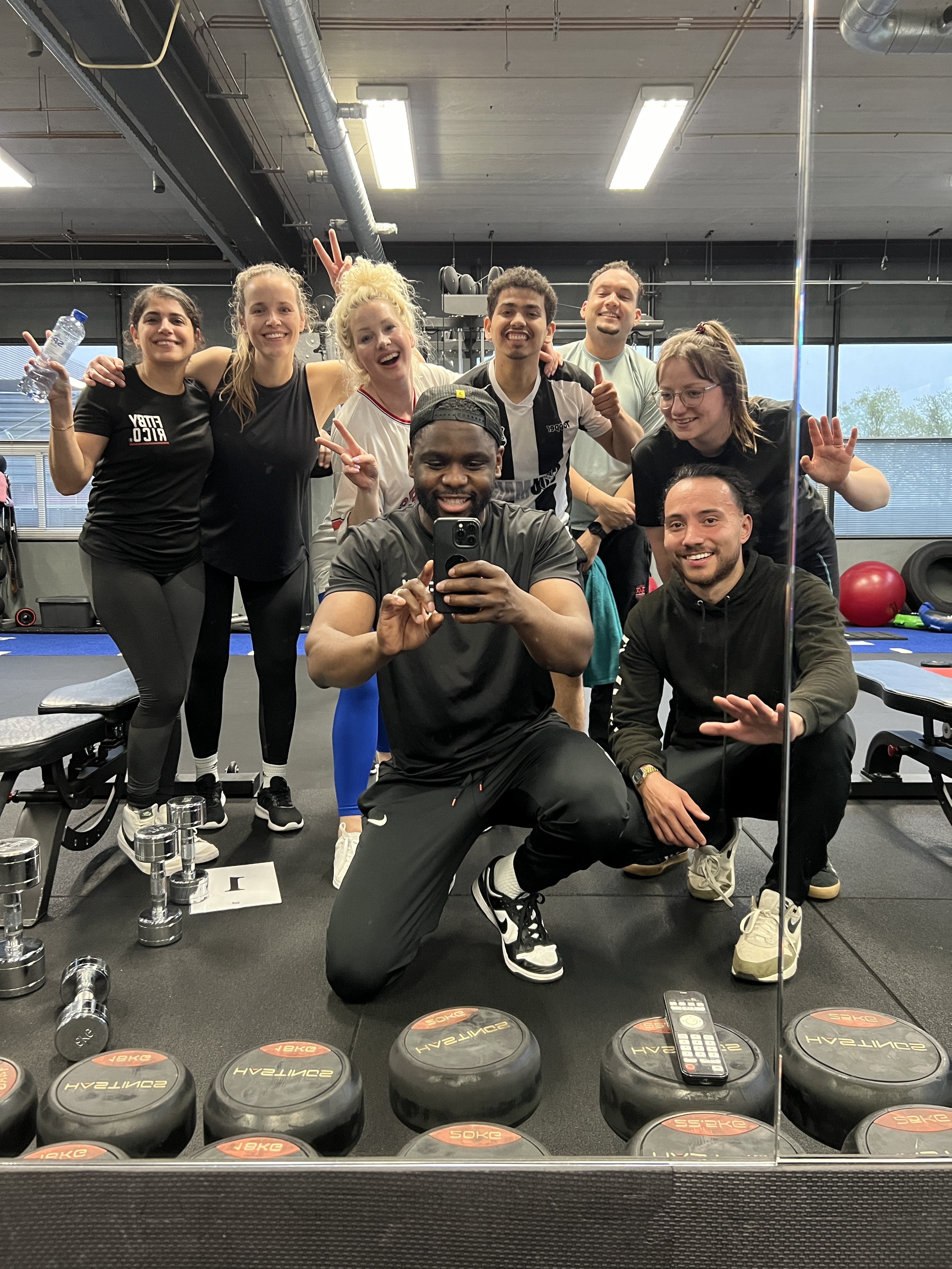 Groep mensen in een sportschool die poseert voor een selfie, omgeven door fitnessapparatuur en gewichten.