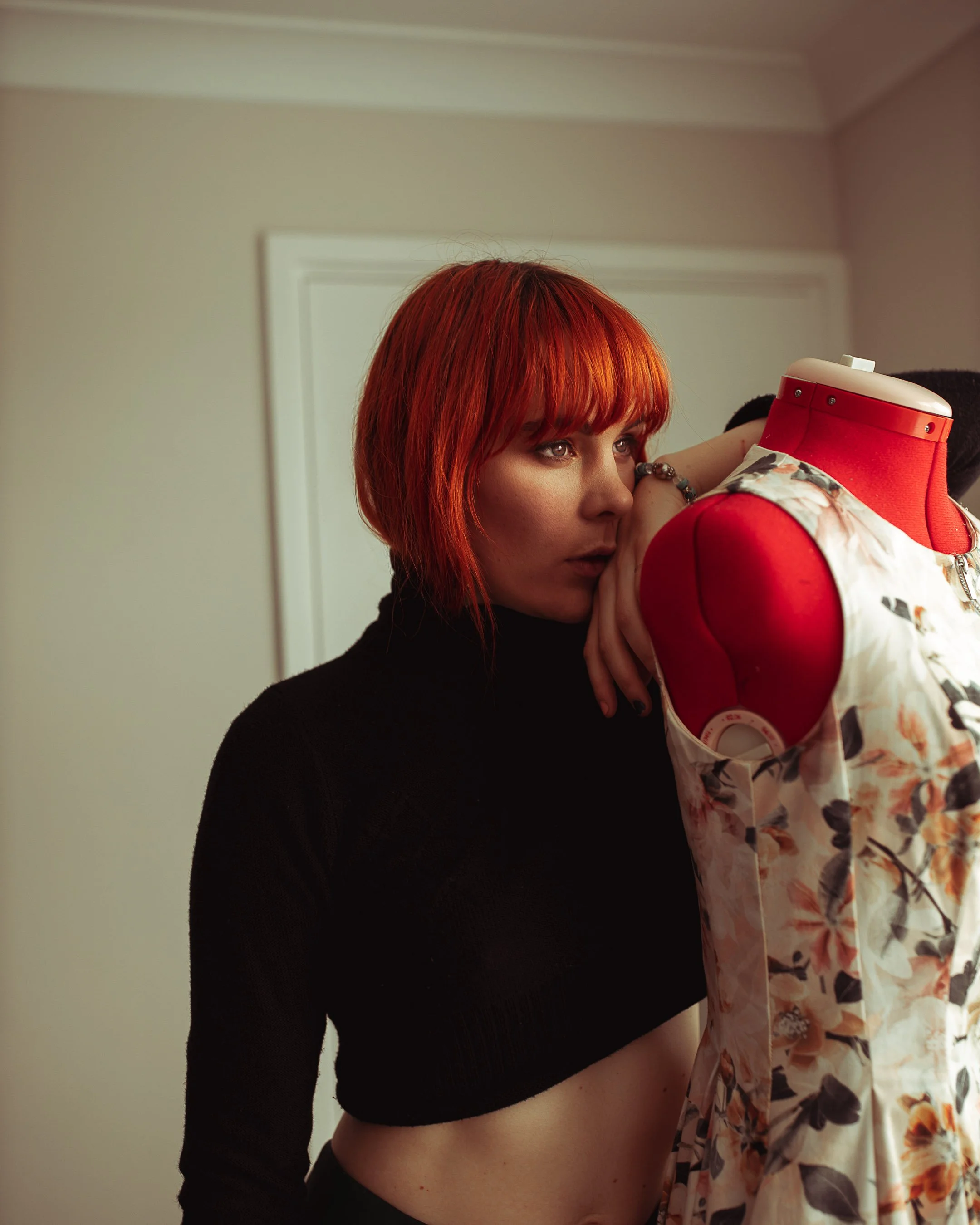 A woman with short red hair leaning on a dress form wearing a floral sleeveless top, inside a room with beige walls.
