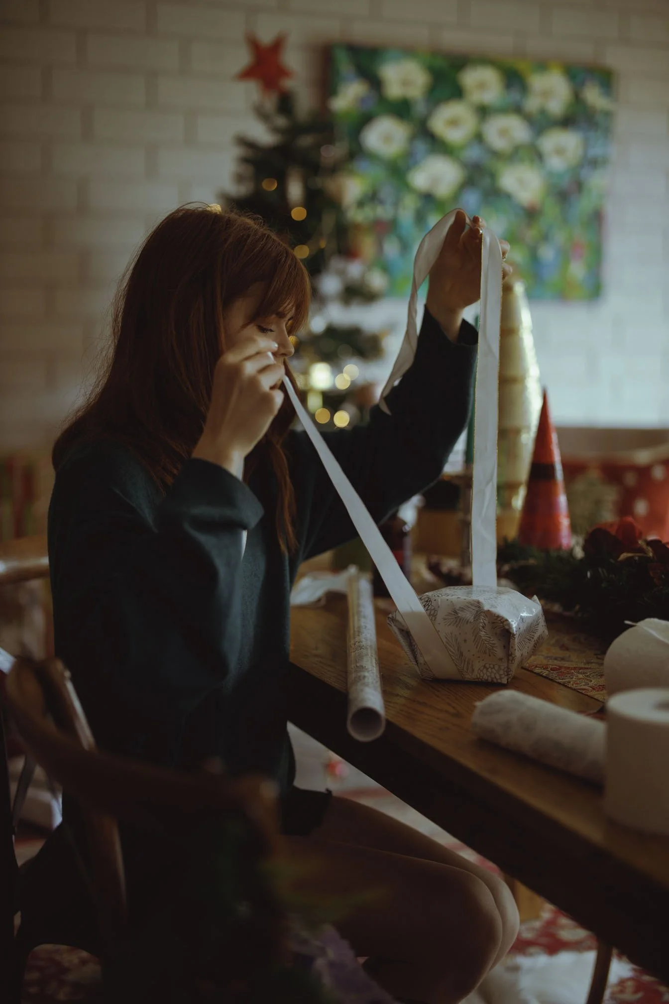 A woman with red hair sitting at a wooden table, wrapping a Christmas gift with festive paper and a white ribbon, with holiday decorations and a Christmas tree in the background.
