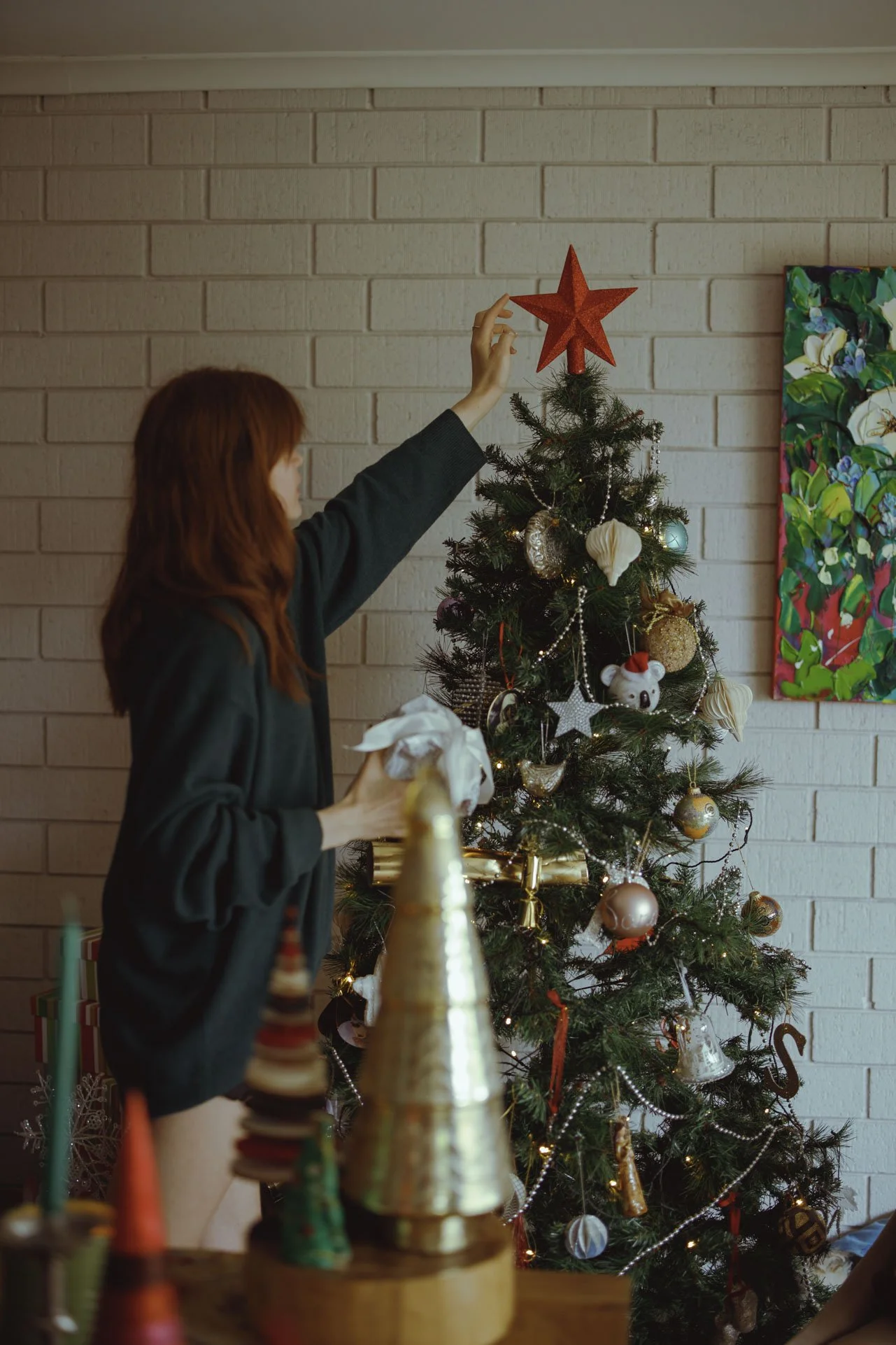 A woman decorating a Christmas tree with a red star on top, standing near a beige brick wall, with colorful ornaments and lights on the tree.